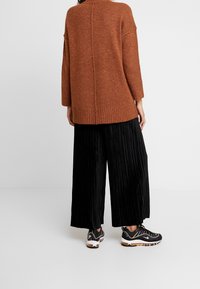 Maglione lavorato a maglia di colore ruggine oversize con orlo a costine, abbinato a pantaloni larghi neri plissettati e sneakers nere con accenti colorati.