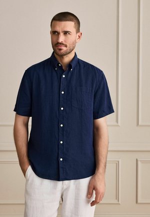 Man with short hair and beard wearing a navy blue short-sleeve button-up shirt and white pants, standing against a beige paneled wall.