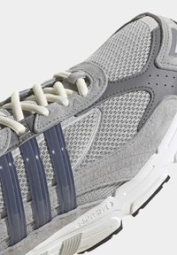 Šedá sportovní bota vyrobená z síťoviny a semiše. Obsahuje tři tmavé pruhy, texturovaný svršek a bílou gumovou podešev.