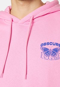 Gros plan sur un sweat à capuche rose avec un motif de papillon bleu et le texte "Obscure Future Fly Like a Butterfly" sur la poitrine, porté par une personne.