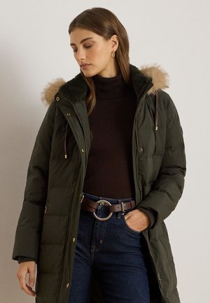 Žena v tmavozelenej prešívanej bunde s kožušinovou kapucňou, čiernom roláku, modrých džínsoch a hnedom opasku, pozerá dole s rukou v vrecku.