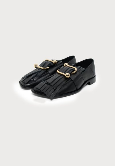 Mocassins en cuir noir avec détails à franges et grandes boucles métalliques dorées sur fond blanc.