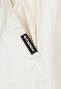 Svetel kremast material z gladko teksturo ima črno oznako Converse blizu šiva, kar poudarja minimalističen dizajn in blagovno znamko.