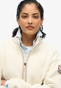 Giacca in pile color panna con collo alto e cerniera frontale, indossata sopra una camicia a righe azzurro chiaro; presenta una toppa sulla manica.