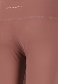 Brune atletiske leggings med en glat tekstur, der har flade syninger og et guld "ENDURANCE" logo nær taljen.