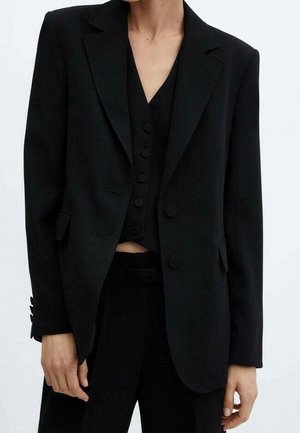 Personne portant un costume noir sur mesure avec un gilet boutonné en dessous, debout devant un fond clair uni.