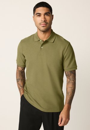 Muž s krátkymi tmavými vlasmi v olivovo zelenom tričku s krátkym rukávom a čiernych nohaviciach, stojaci s jednou rukou v kapse.