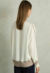 Lätt, krämfärgad jacka med långa ärmar och ribbad beige nederkant. Har en slät yta och subtila horisontella randdetaljer.