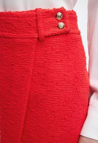 Κοντινό πλάνο κόκκινης υφασμάτινης φούστας με ψηλή μέση και δύο χρυσά διακοσμητικά κουμπιά, φορεμένη με λευκό μπλουζάκι μακρυμάνικο.