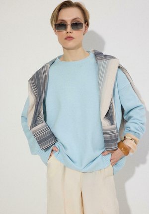 Personne portant un pull bleu clair et un pantalon crème, veste rayée drapée sur les épaules, lunettes de soleil et larges bracelets au poignet.