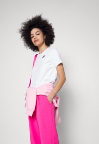 T-shirt blanc Nike avec un logo noir, associé à un pantalon de survêtement rose vif. Sweat à capuche rose noué autour de la taille. Texture de tissu lisse.