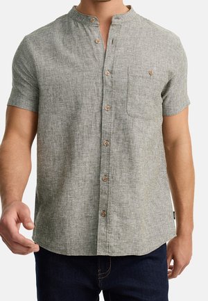 Mężczyzna noszący jasnoszarą, teksturowaną koszulę na guziki z krótkim rękawem, kołnierzykiem typu mandarynka i kieszenią na piersi, w połączeniu z ciemnoniebieskimi spodniami.