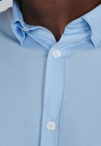 Jasnoniebieska koszula na guzik z teksturowanej tkaniny, z kołnierzykiem w kształcie punktu i białymi guzikami z przodu. Zbliżenie na dekolt i szczegóły tkaniny.