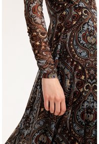 Nahaufnahme einer Hand einer Person, die ein langärmeliges Kleid mit komplizierten Paisleymustern in braunen, blauen und roten Farbtönen trägt.