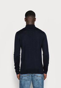 Pull en maille bleu marine foncé avec un col montant, des manches longues et des poignets côtelés. Porté avec un jean bleu délavé, présentant un motif subtil.