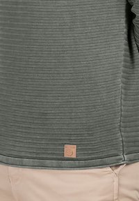 Grau gestreifte Shirt aus strukturiertem Stoff, mit einem braunen Logopatch am Saum und einer lässigen Passform. Akzente sind subtile Steppdetails.