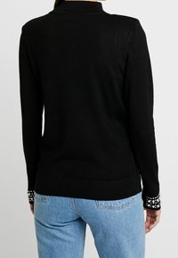 Kvinna iklädd en figurnära svart långärmad tröja med pärldekorationer på ärmsluten och ljusblå jeans, sedd bakifrån.