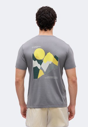 Szürke pólót viselő férfi, amelyen sárga, fehér és zöld absztrakt hegy- és napmotívum szerepel, hátul kalandról szóló szöveggel.