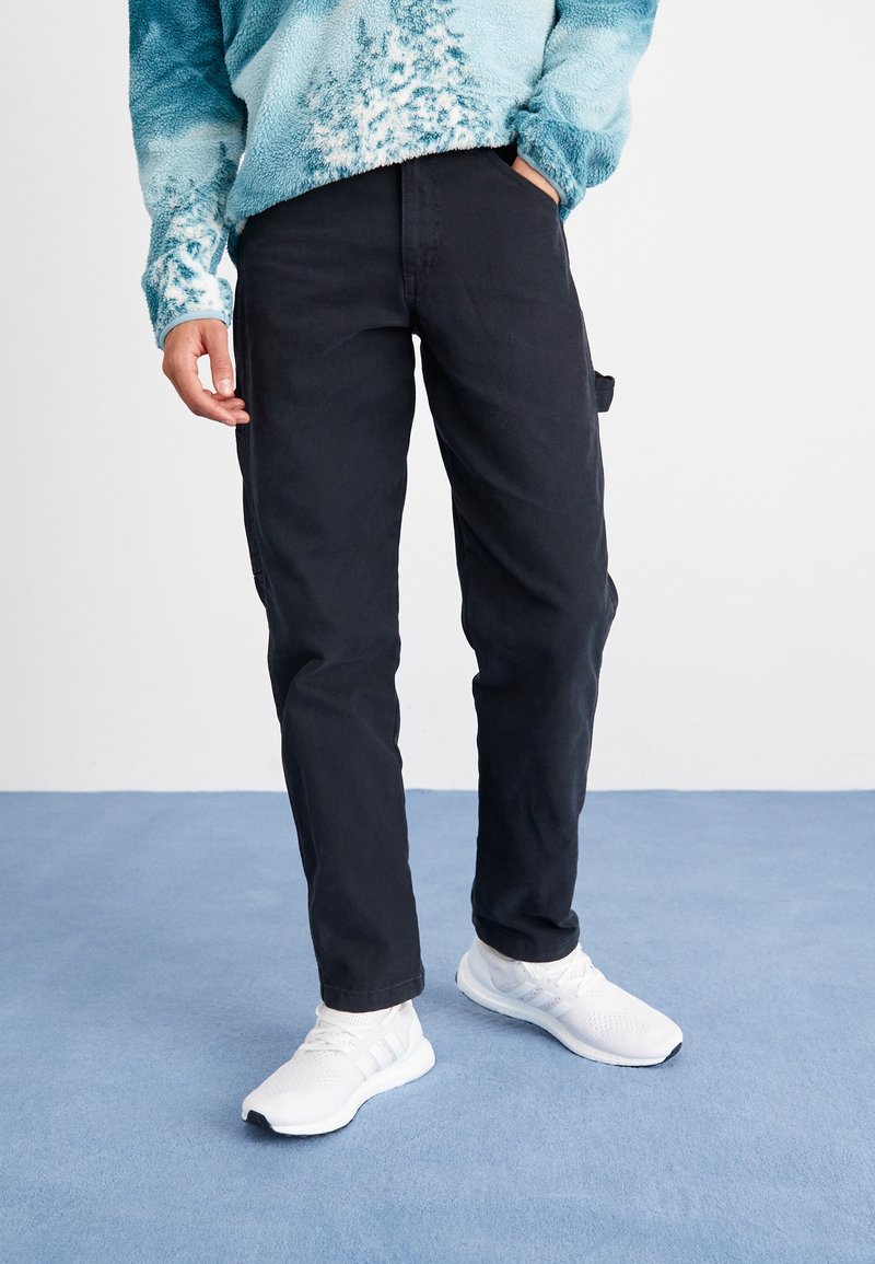 Personne portant un pantalon noir, des baskets blanches et un pull polaire bleu à motifs, debout sur un sol moquetté bleu.