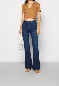 Brun, kortärmad topp med V-ringning; mörkblå utsvängda jeans; beige spännbandssandaler; ljusblå handväska med axelrem.