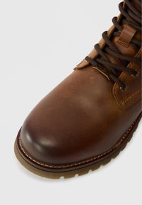 Brūna ādas zābaks ar tumši brūnām auklām, izstrādāta šuvuma un izturīgu zoli uz balta fona.