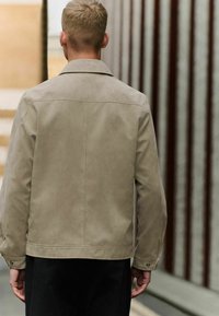 Chaqueta de ante beige con cuello, bolsillos laterales y puños abotonados, llevada sobre un atuendo oscuro, vista desde la parte posterior.