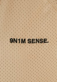 Šviesiai rudos perforuotos medžiagos, puoštos juodu siuvinėtu tekstu "9N1M SENSE." Dizainas yra lengvas, turi kvėpuojančią tekstūrą ir nuoseklų raštą.