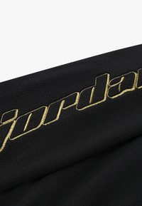 Tessuto nero con una superficie texturizzata che presenta il logo "jordan" ricamato in oro, posizionato diagonalmente nell'immagine.