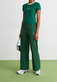 Camiseta verde ajustada con el logo "Nike", combinada con pantalones verdes holgados y zapatillas blancas. Incluye un collar de cadena plateada y un pequeño bolso verde.