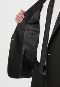 Czarna marynarka o gładkiej zewnętrznej fakturze, z czarną satynową podszewką, z pojedynczym zapięciem na guzik i detalem wewnętrznej kieszeni.