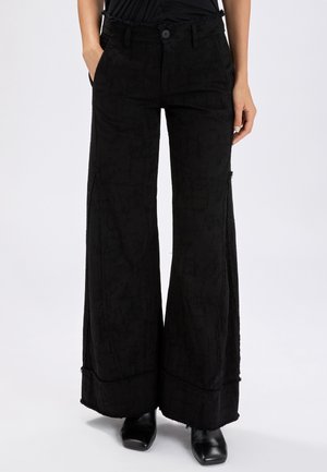 Pantaloni neri a gamba larga con tessuto strutturato e bordi sfrangiati, indossati con scarpe nere a punta e una maglietta nera infilata dentro.