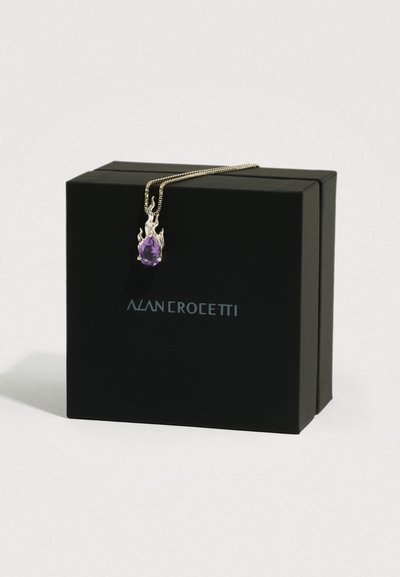 Złoty naszyjnik z zawieszką w kształcie płomienia, trzymającą fioletowy kamień w kształcie łzy, wyeksponowany na czarnym pudełku Alan Crocetti.