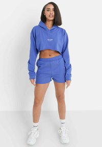 Afgesneden blauwe hoodie en bijpassende shorts van zacht materiaal. De hoodie heeft een logo; de shorts hebben een elastische tailleband. Witte sneakers en sokken.