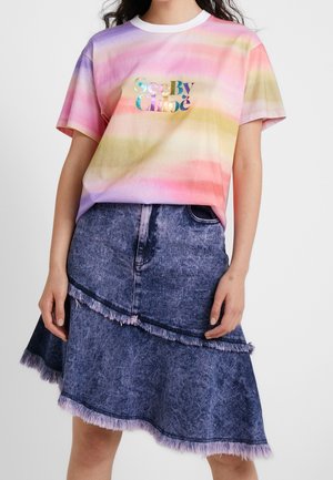 Frau trägt ein pastellfarbenes Batik-T-Shirt mit mehrfarbigem Text und einen blauen asymmetrischen Jeansrock mit ausgefransten Kanten.