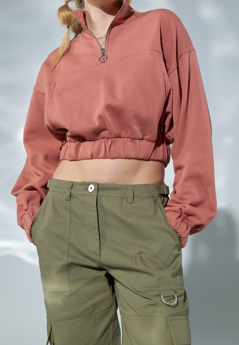 Personne portant un sweat rose poudré court à manches longues et un pantalon cargo vert olive, les mains dans les poches, debout devant un fond clair.