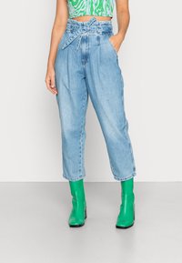 Calças de ganga de cintura alta azul claro com um laço na cintura e bolsos laterais, combinadas com botas de tornozelo verdes brilhantes.