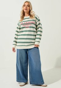 Gestreiftes Langarmhemd in Creme mit grüner und roter Schrift, kombiniert mit weiten blauen Jeans und beigen Slippern.