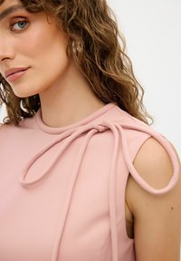 Blouse rose avec un col rond, des nouettes aux épaules et des découpes. Le tissu semble lisse et fluide.