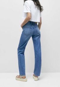 Mujer con jeans rectos de tiro alto azul, camiseta corta blanca y sandalias de plataforma beige apoyada contra una pared lisa.