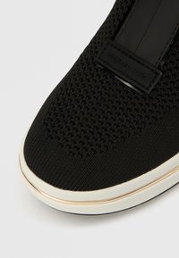 Sneaker in maglia nera con tomaia testurizzata, suola in gomma bianca e striscia decorativa dorata. Include un'etichetta del logo in pelle nera.