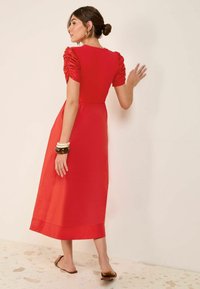 Robe midi rouge avec des manches courtes et ballons, texture lisse, détails froncés aux épaules et ourlet droit. Portée avec des sandales marron.
