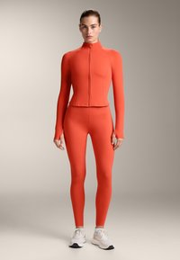 Oranžový športový odev pozostávajúci z priliehavého topu s dlhým rukávom a vysokým golierom a zodpovedajúcich legín, vyrobený z pružného a hladkého materiálu.