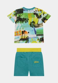 T-shirt z wielokolorowym wzorem kolażu przedstawiającym palmy; zestawiony z szortami w odcieniu teal i żółtym elastycznym pasem.