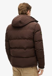 Chaqueta de plumas marrón con capucha, que presenta un diseño acolchado, puños elásticos y el logo "Superdry JPN" bordado en la parte posterior.