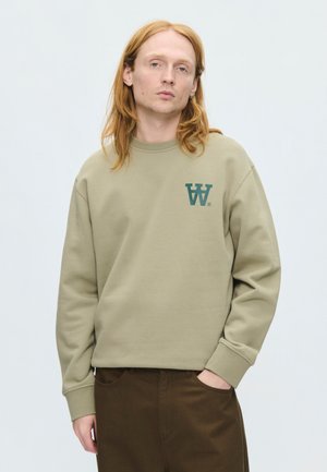 Person mit langen roten Haaren trägt ein hellbraunes Sweatshirt mit blauem Logo und dunkelbraune Hose, steht mit einer Hand in der Tasche.