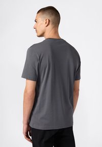 Mę�żczyzna z krótkimi włosami, ubrany w prostą ciemnoszaro t-shirt z krótkim rękawem oraz czarne spodnie, stojący bokiem na białym tle.