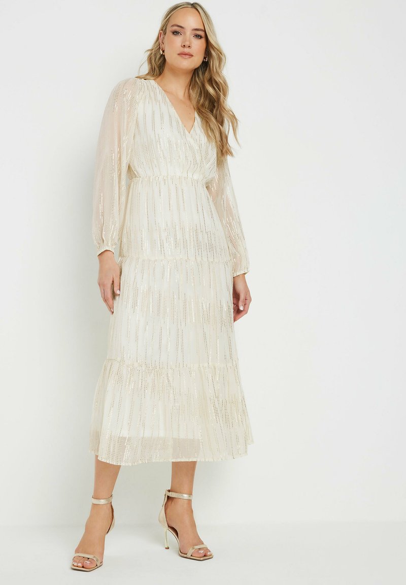 Vestido maxi de capa en crema con brillo, mangas largas translúcidas y escote en v. Combinado con sandalias de tacón con tira metálica en el tobillo.