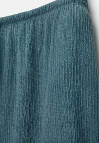 Teal strukturerad tyg med ett vertikalt ribbat mönster, med en gummiband i midjan. Närbild av material och design detaljer.