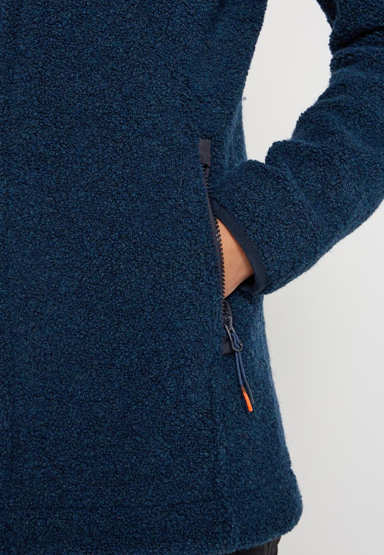 Hand in einer navyblauen, strukturierten Fleecejackentasche mit sichtbarem Reißverschluss und orangefarbenem Zipper an einem schlichten Hintergrund.