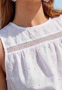 White cotton blouse featuring floral embroidery and openwork detailing across the chest. Smooth texture with a rounded neckline and sleeveless design.